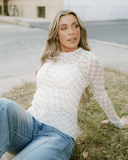 Model wearing a khaki sheer checkered top with long sleeves, sitting on the grass in casual blue jeans.