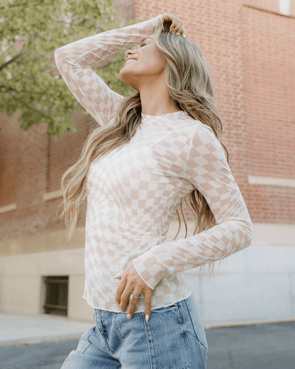 Woman wearing a khaki sheer checkered top posing outdoors with a playful expression against a brick wall.