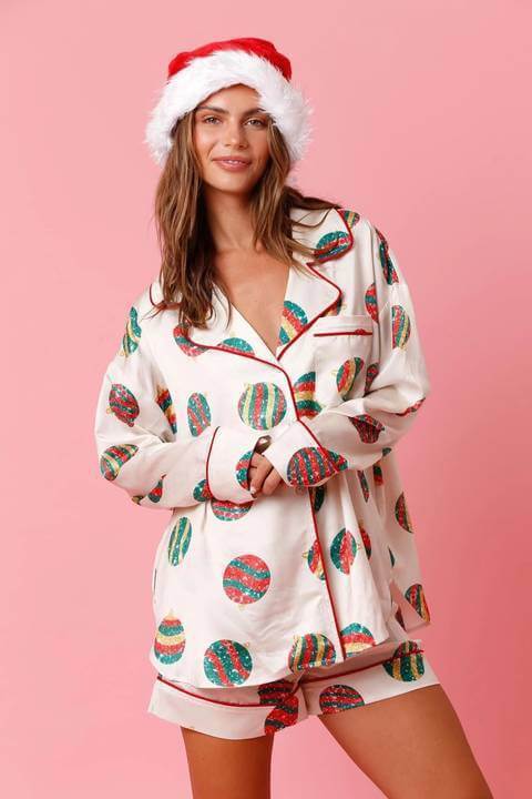Smiling woman in a Santa hat wearing a women's satin santa pajama set with Christmas ornaments on a pink background.